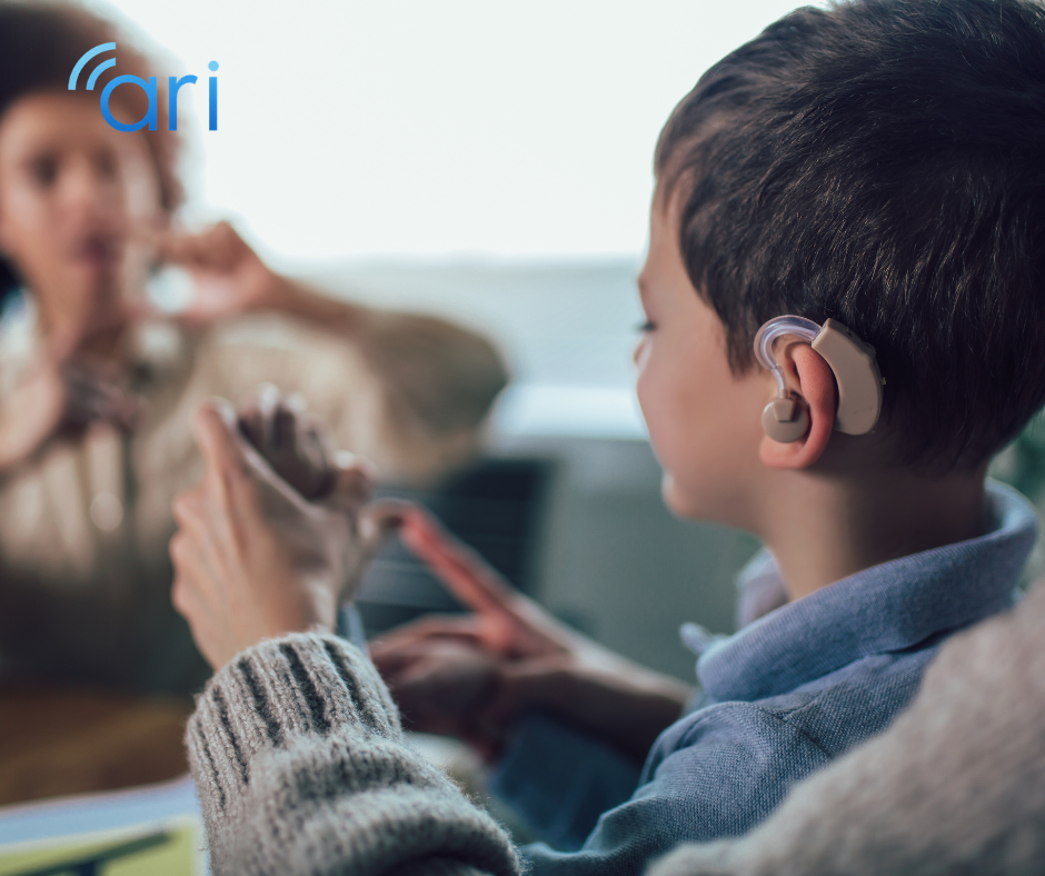 Young child wearing a cochlear implant using sign language while facing an adult in a home environment, demonstrating multimodal communication.