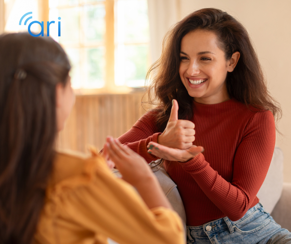 A smiling woman using sign language while sitting on a couch and communicating face to face with a young girl in a bright home setting.