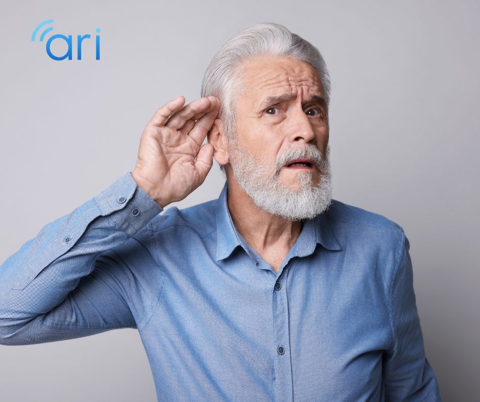 An older man gestures toward his ear, indicating difficulty hearing or a desire to hear more clearly. The image represents hearing loss and the importance of communication access in healthcare settings.