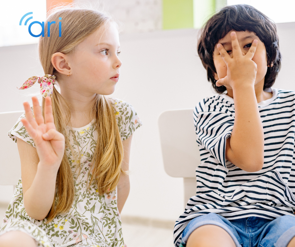 Two young children sitting side by side in a classroom setting using hand gestures that resemble sign language, demonstrating early communication and visual language development.