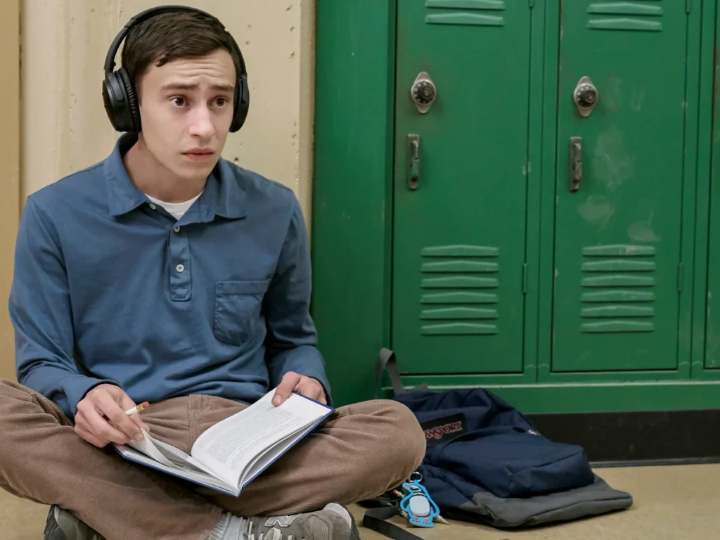 Scene from the TV show Atypical showing a teenage boy wearing headphones sitting on a school hallway floor reading a book beside green lockers.