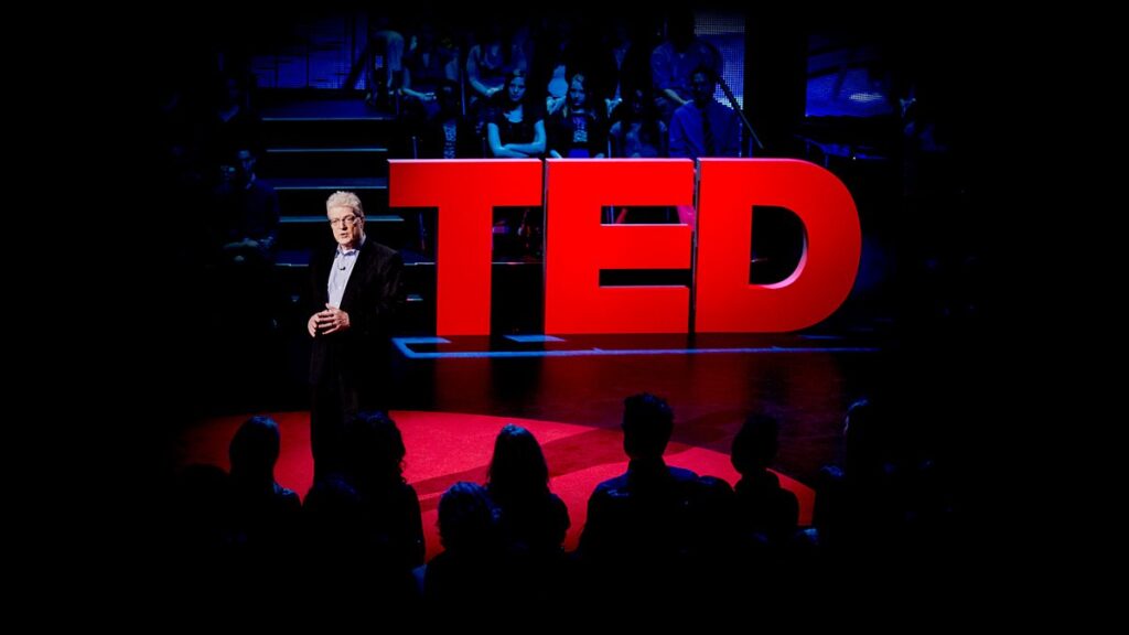 Speaker standing on a TED stage in front of large red TED letters, delivering a talk to a seated audience in a dark auditorium.